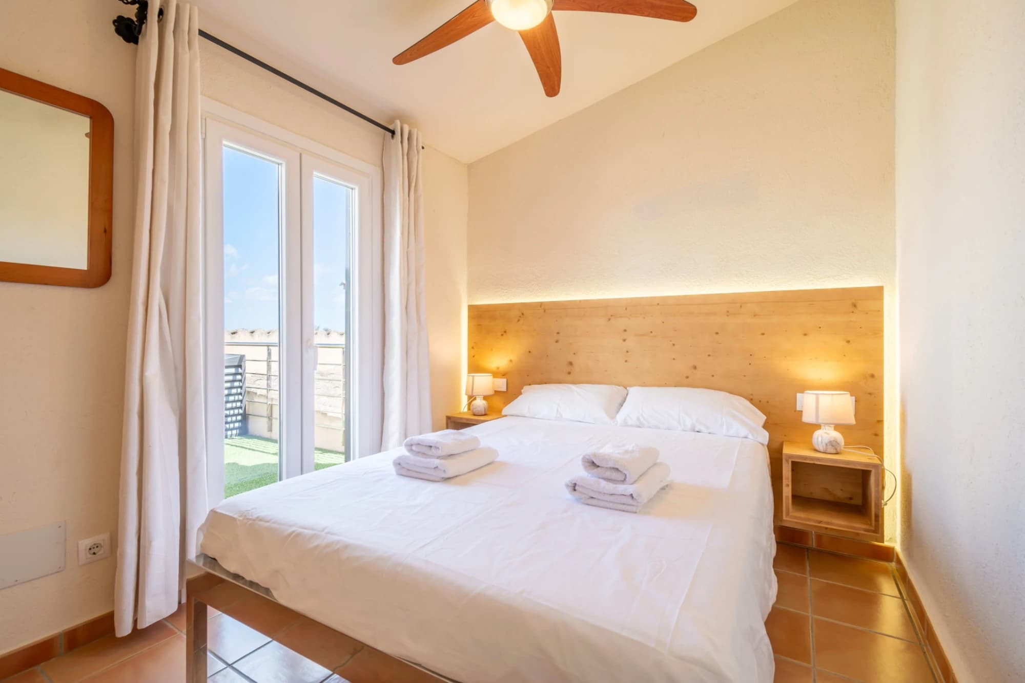 A hotel room with a bed, a ceiling fan, a nightstand with a lamp, and a view of the outdoors.